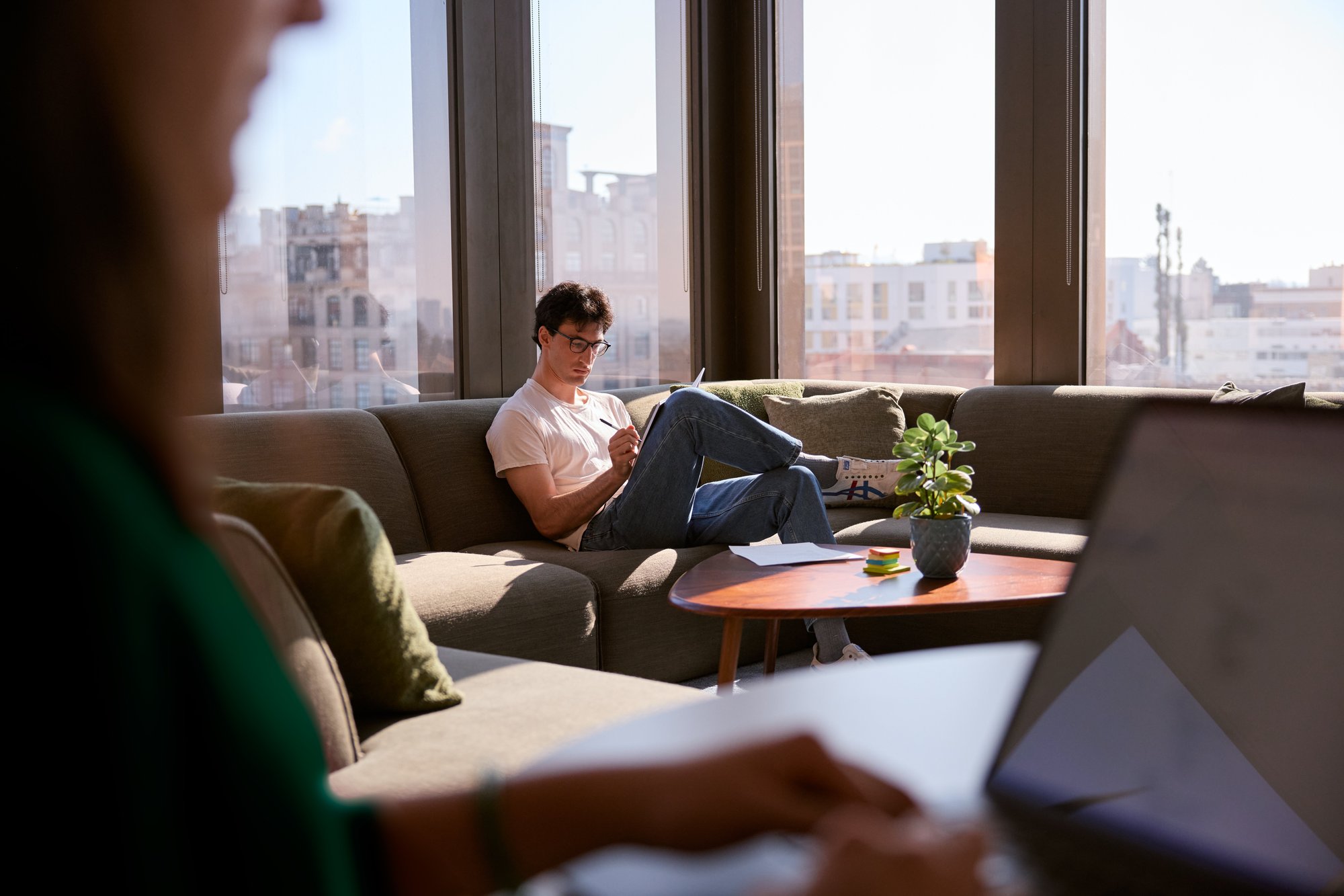 One solo figure writing with pen and paper on a couch in front of large windows.