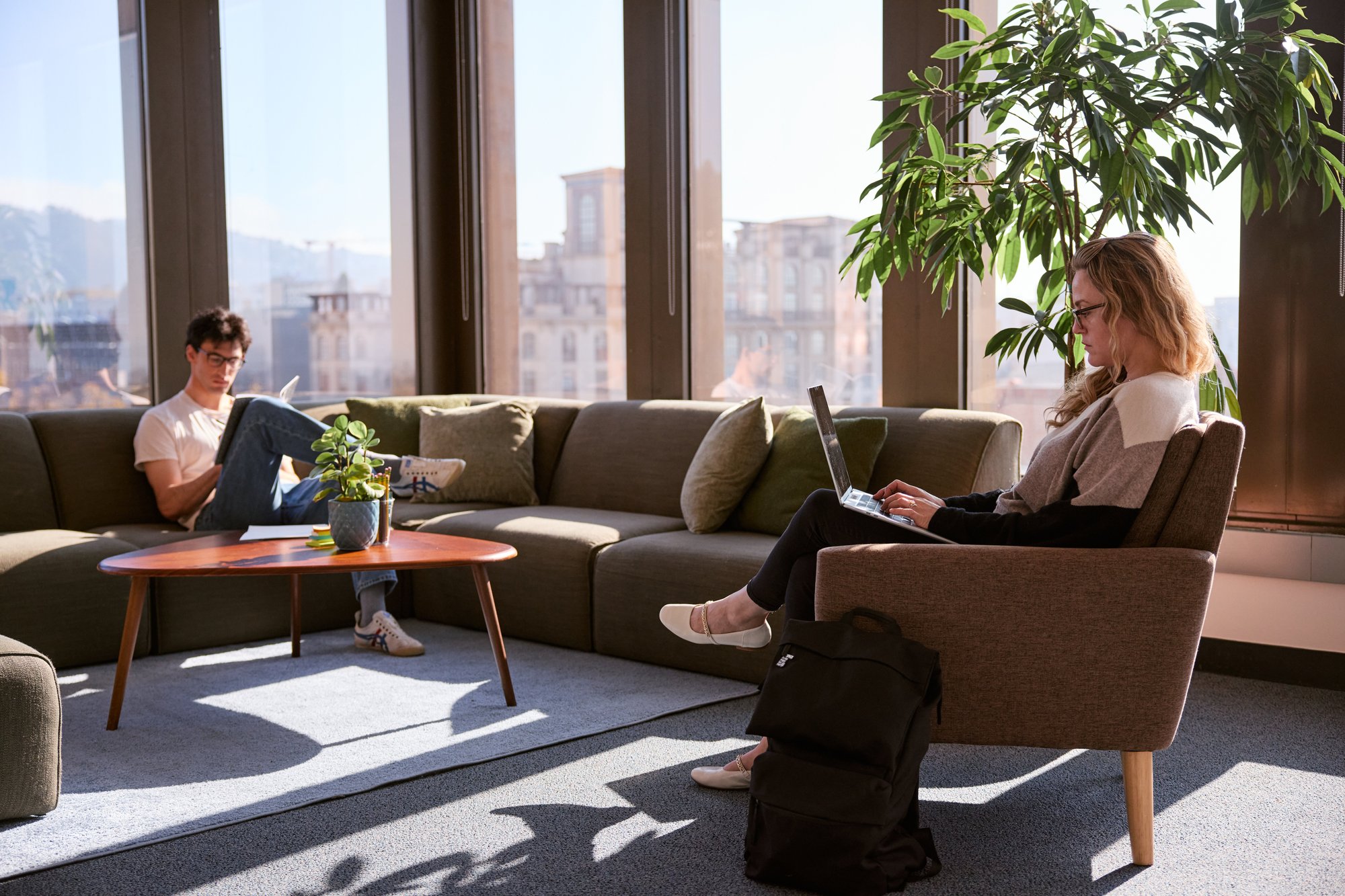 Two people focused on their work. One sits in a chair and the other on a couch in front of large windows.