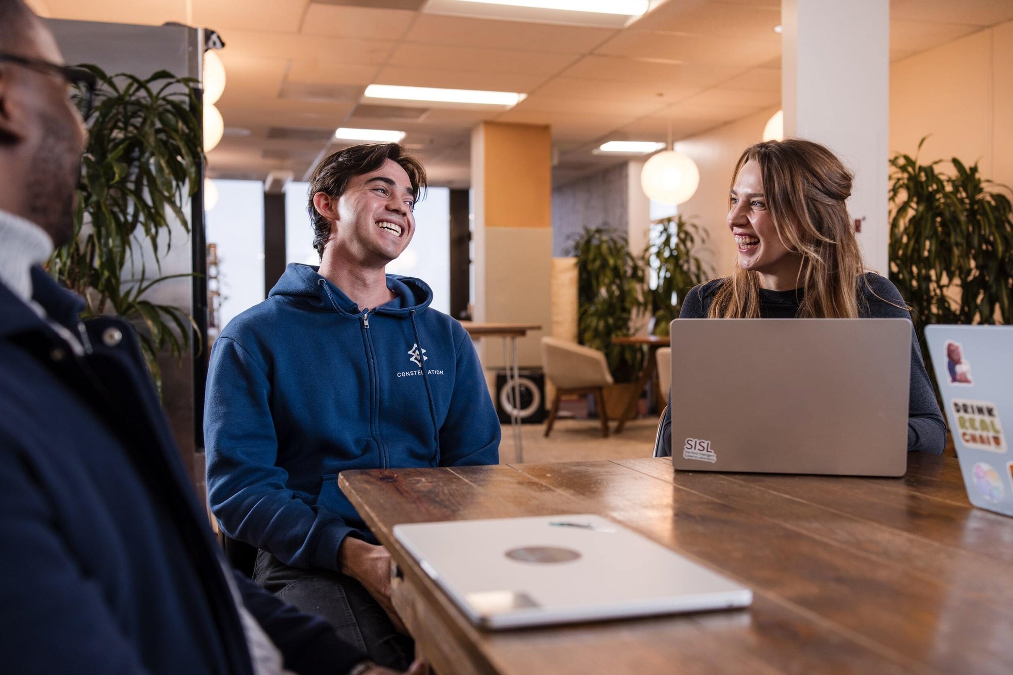 Person wears Constellation hoodie. They are laughing and smiling while two other people at the table smile back. 