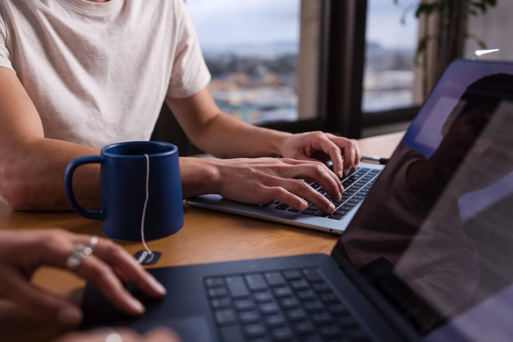 Close up image of two people typing on laptops with a cup of tea on the table.