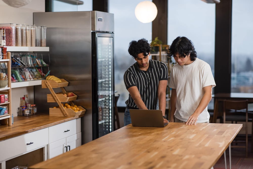 Two people standing at a table and working on a shared laptop.